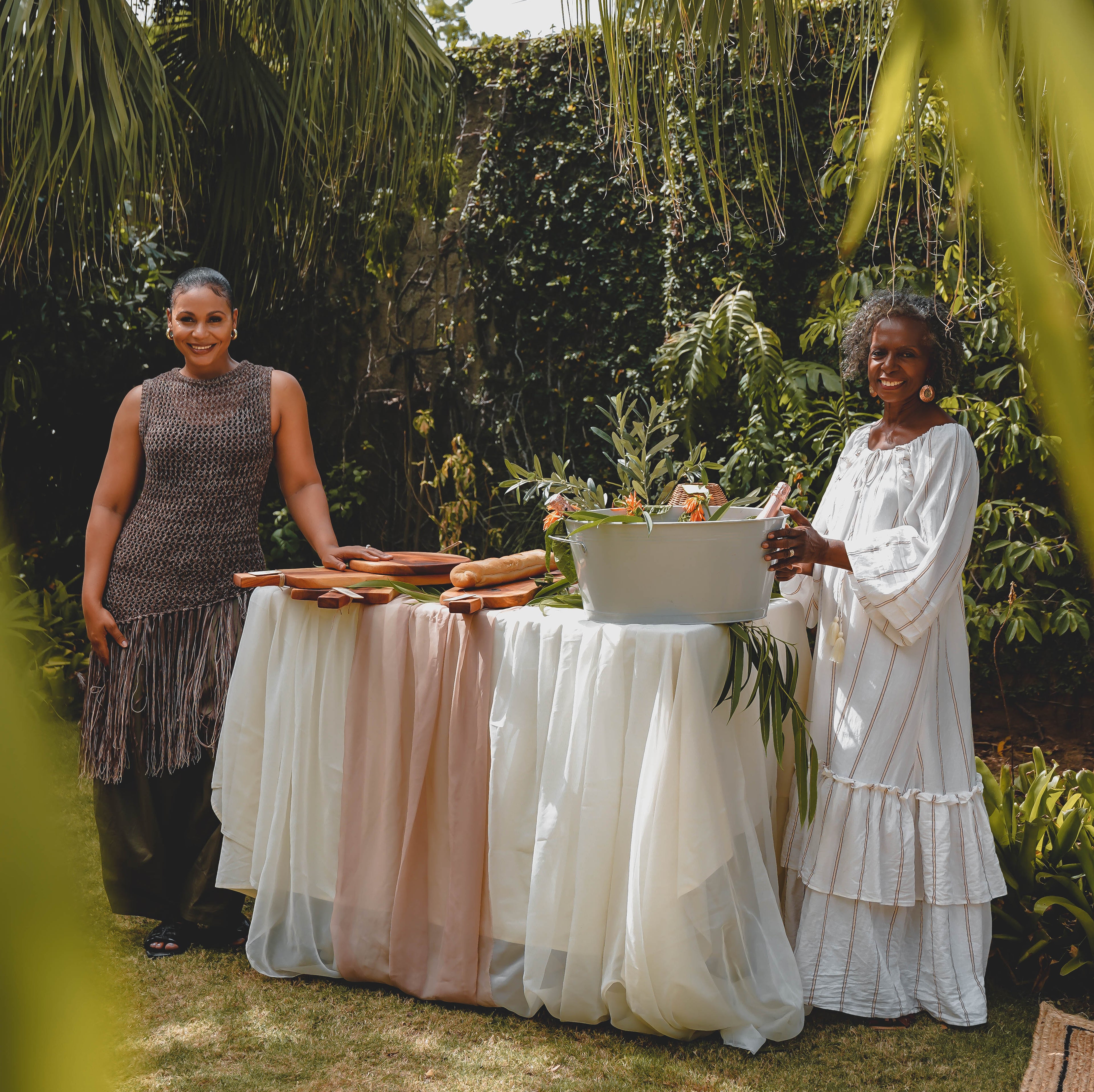 Two women standing outdoors at a decorated table draped in white and blush pink fabric, with palm leaves framing the scene and greenery in the background.